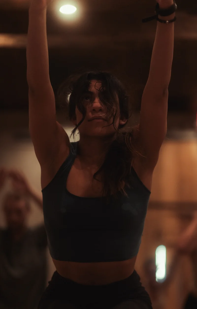 A woman practices chair pose with raised arms and sweat across her skin in the warm studio.